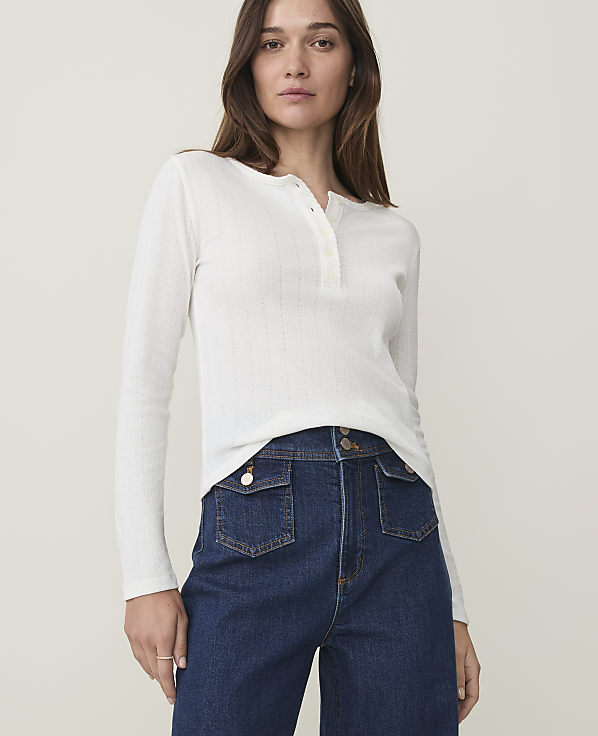 A woman in a white henley shirt and dark wash jeans stands against a plain, light background.