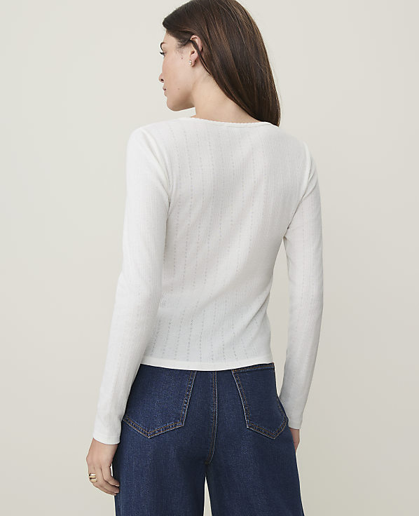 A woman in a white long-sleeved shirt and blue jeans stands against a plain, light-colored background.