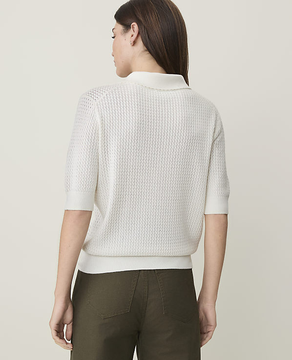 A woman in a white knit polo shirt and olive green pants stands against a plain white background.