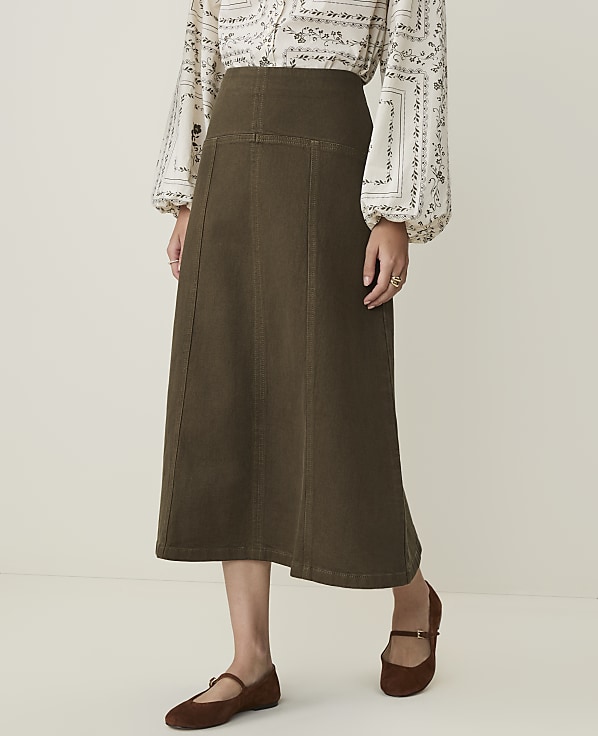 A woman in a patterned blouse and a long olive-green skirt stands against a plain, light-colored background.