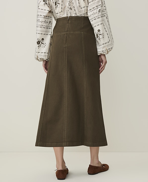 A person wearing a patterned blouse and a long olive-green skirt stands against a plain, light-colored background.