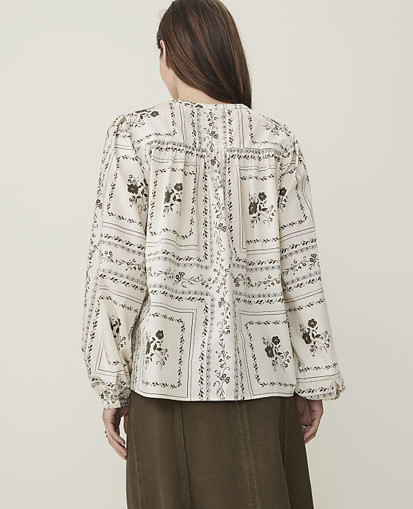 A woman in a patterned blouse and olive skirt stands against a plain, light background.