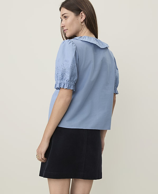 A woman in a blue ruffled blouse and dark skirt stands against a plain, light background.