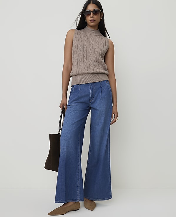 A woman in a sleeveless cable-knit sweater and wide-leg jeans stands against a plain white background, holding a dark tote bag.