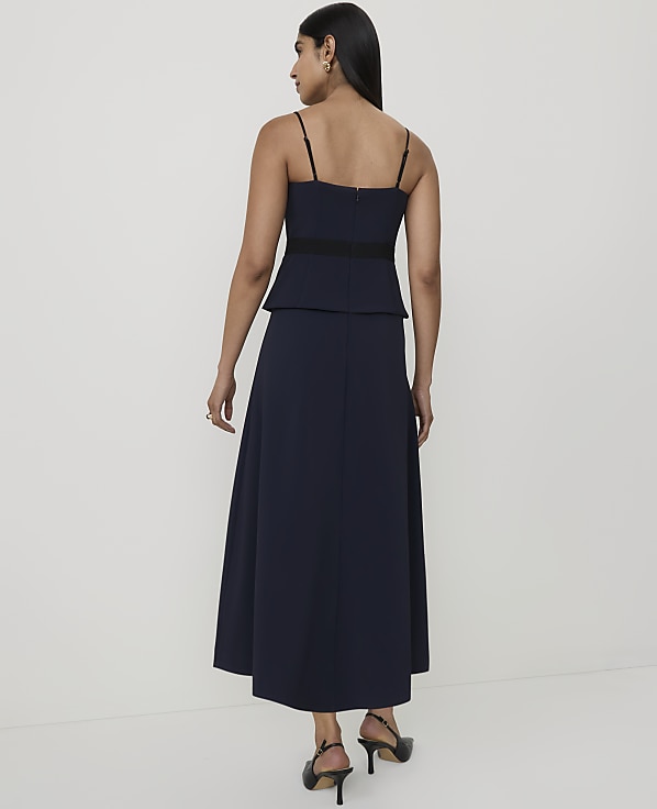 A woman in a navy blue dress with spaghetti straps and a black waistband stands against a plain white background.