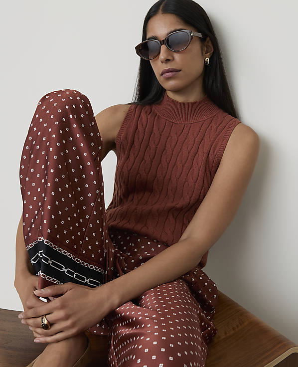 A woman in a rust-colored cable-knit sleeveless top and patterned wide-leg pants poses against a plain background.