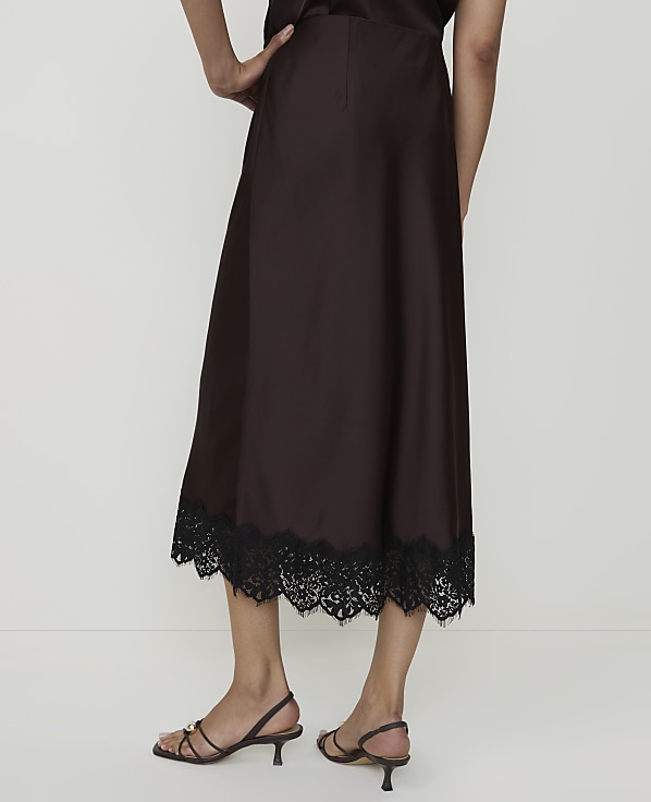 A person stands in front of a plain white background, wearing a brown satin skirt with a black lace hem and strappy sandals.