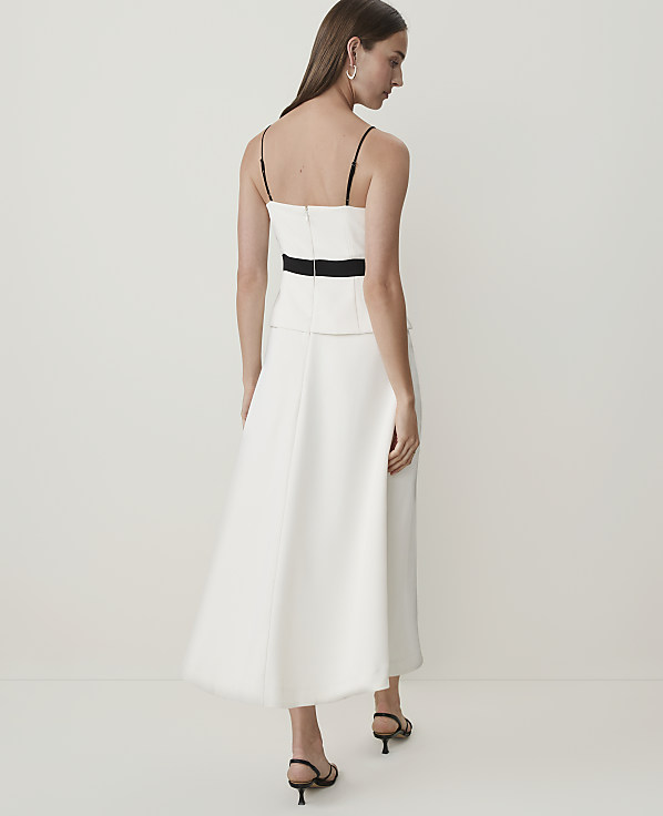 A woman in a white dress with a black belt stands against a plain white background.