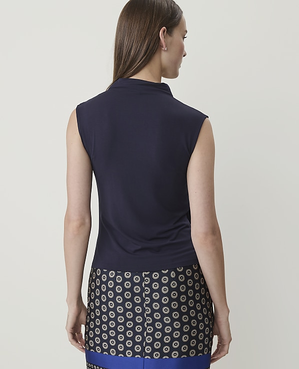 A woman in a navy sleeveless top and a patterned skirt stands against a plain white background.