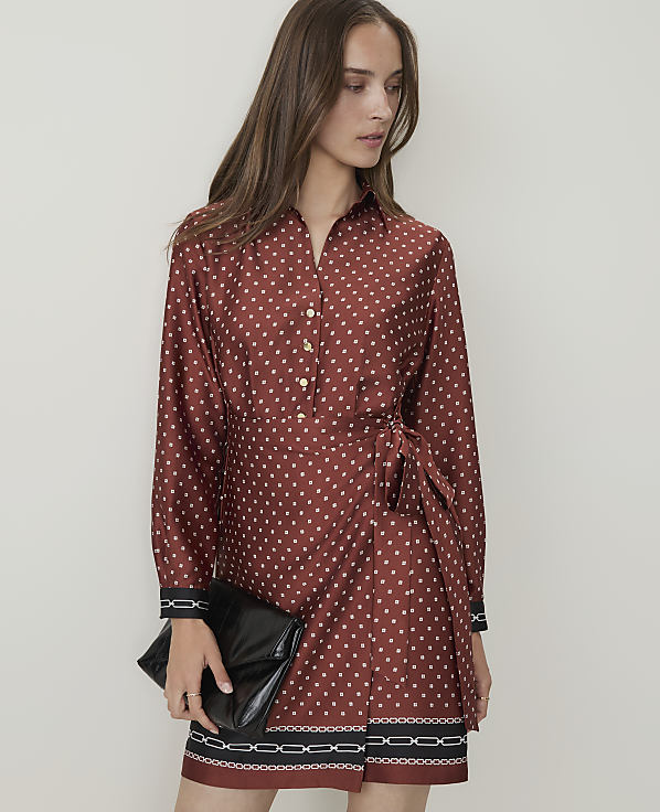 A woman in a patterned rust-colored dress with a black clutch stands against a plain white background.