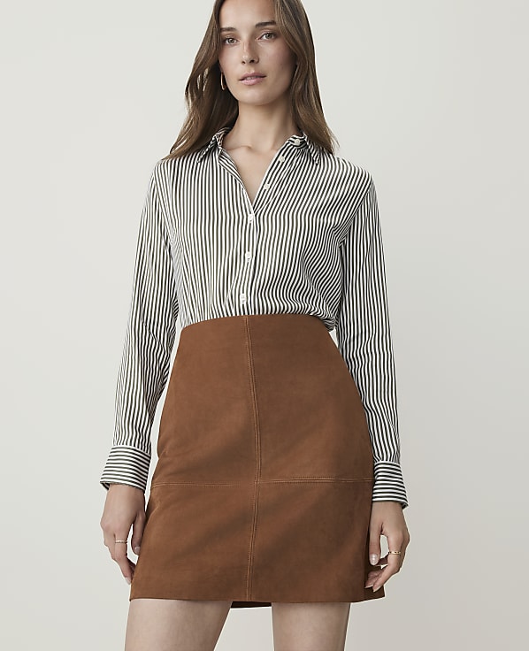 A woman in a striped shirt and brown suede skirt stands against a plain white background.