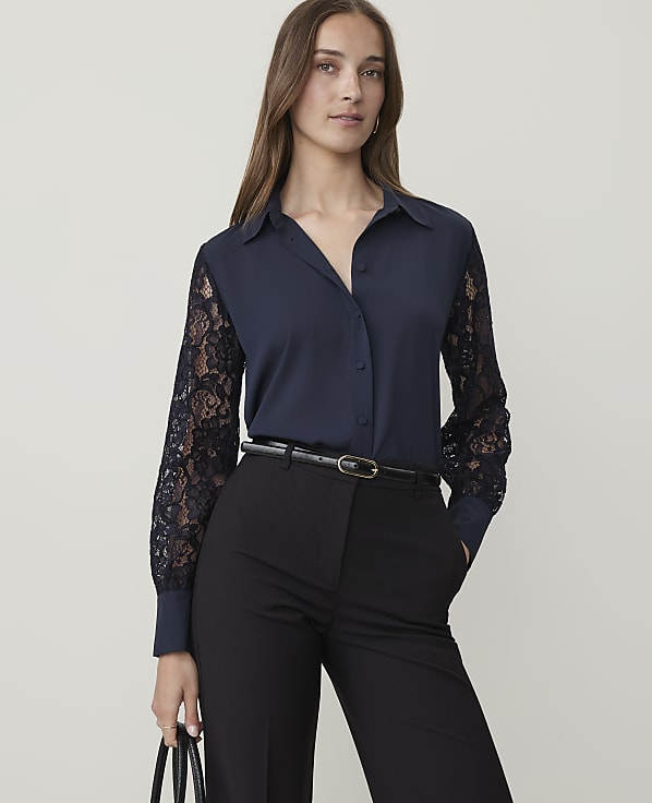 A woman in a navy blouse with lace sleeves and black pants stands against a plain background.