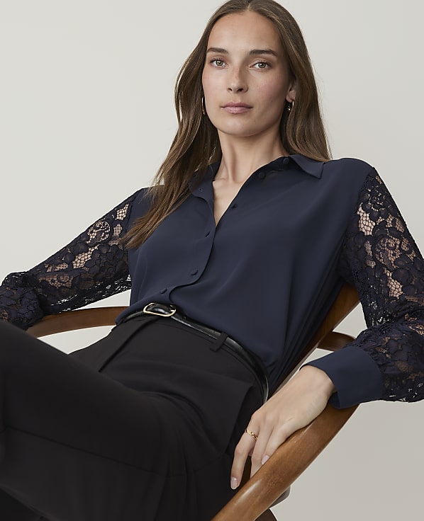 A woman in a navy blue blouse with lace sleeves and black pants sits in a chair against a plain background.