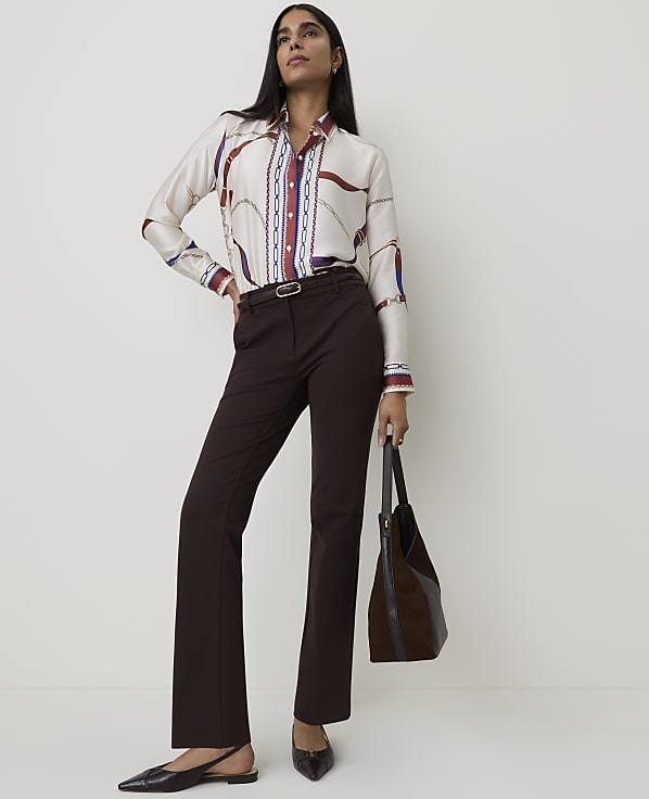 A woman in a patterned blouse and brown pants stands against a plain white background, holding a dark brown bag.