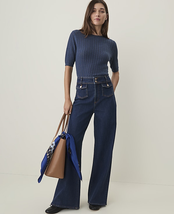 A woman in a blue ribbed sweater and dark wash wide-leg jeans stands holding a brown tote bag with a blue scarf, against a plain, light background.