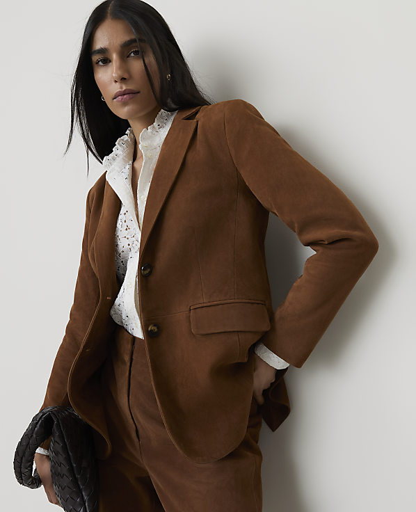 A woman in a brown blazer and pants stands against a plain wall, holding a dark clutch.