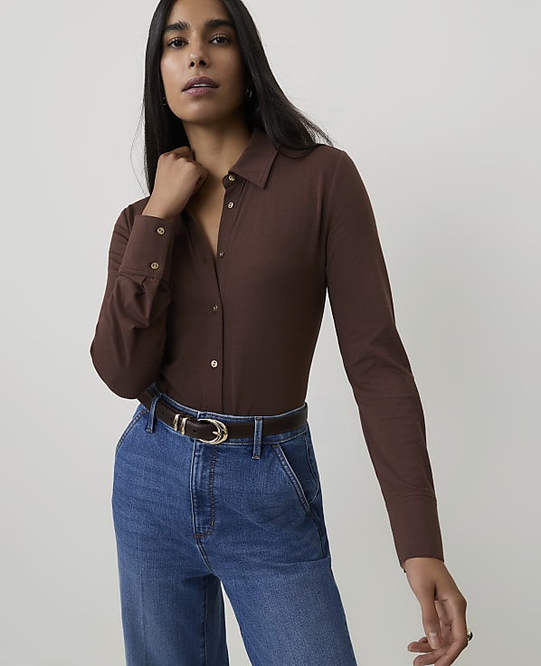 A woman in a brown button-up shirt and blue jeans stands against a plain white background.