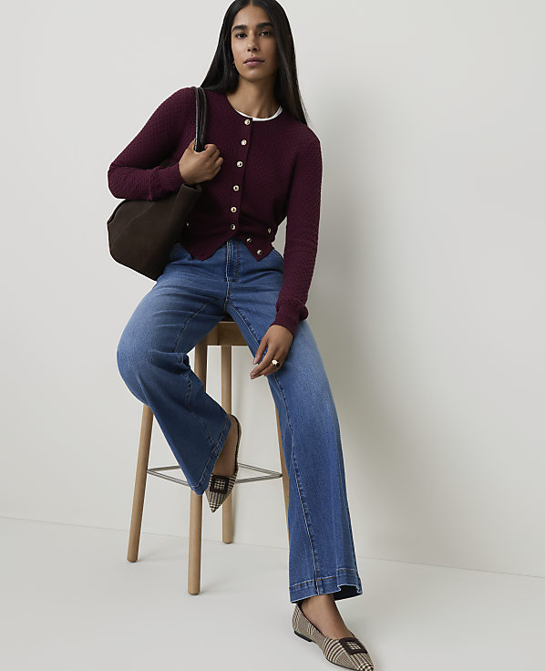A woman in a burgundy cardigan and wide-leg jeans sits on a stool with a brown tote bag, against a plain white background.
