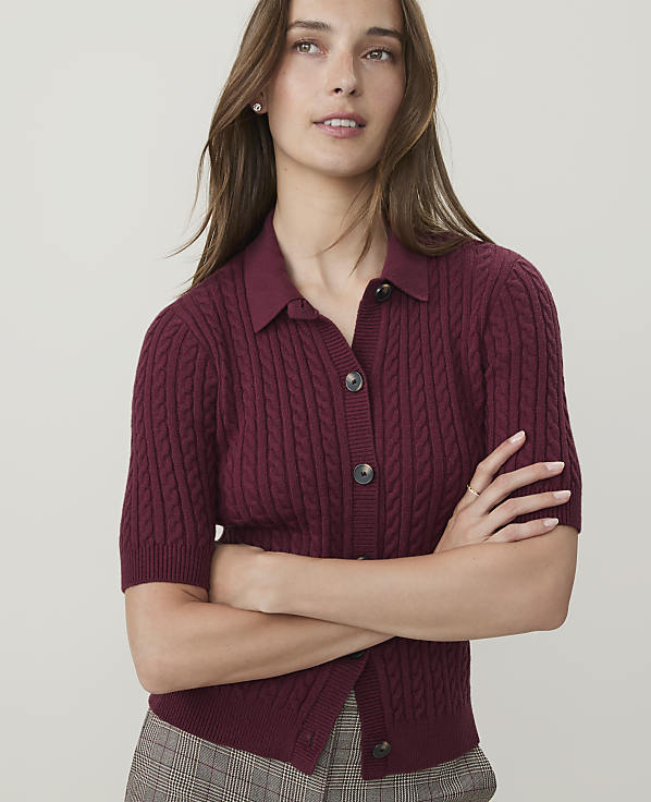 A woman in a burgundy cable-knit polo shirt with her arms crossed stands against a plain white background.