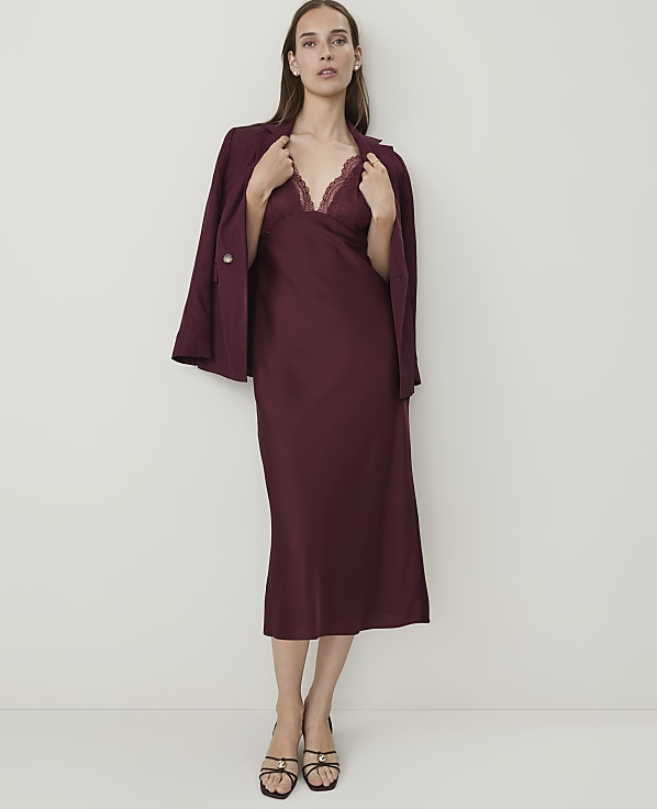 A woman in a burgundy slip dress and blazer stands against a plain white background.