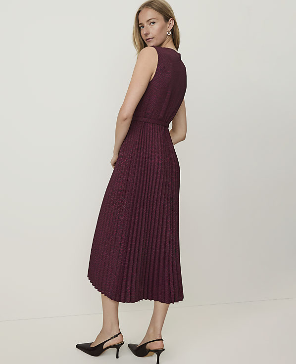 A woman in a burgundy pleated dress and black heels stands against a plain white background.