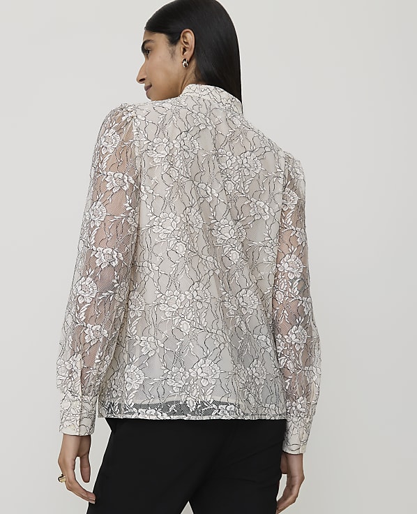 A woman in a white floral lace blouse stands against a plain background.