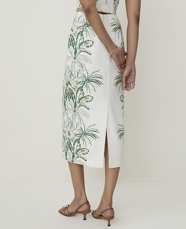 A person stands in the foreground wearing a white skirt with a green tropical leaf pattern, while the background is a plain, light-colored wall.