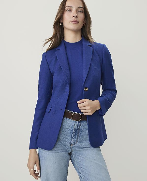 A woman in a blue blazer and jeans stands against a plain, light background.