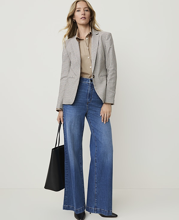 A woman in a patterned blazer and wide-leg jeans stands against a plain, light background, holding a black tote bag.
