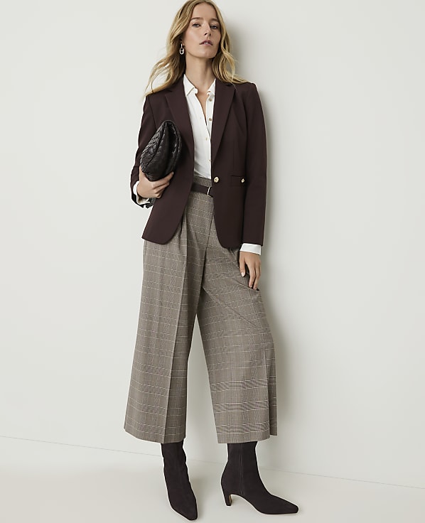 A woman in a brown blazer and plaid culottes stands against a plain white background, holding a black clutch.
