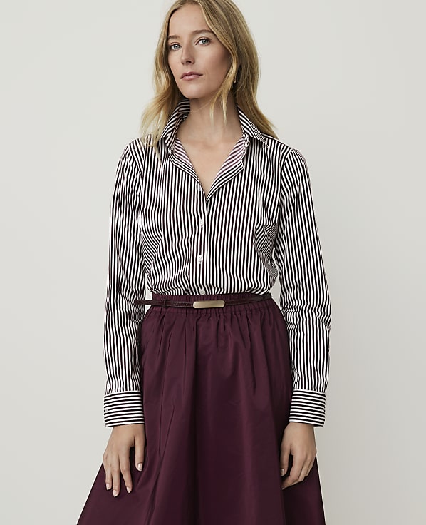 A woman in a striped shirt and burgundy skirt stands against a plain white background.