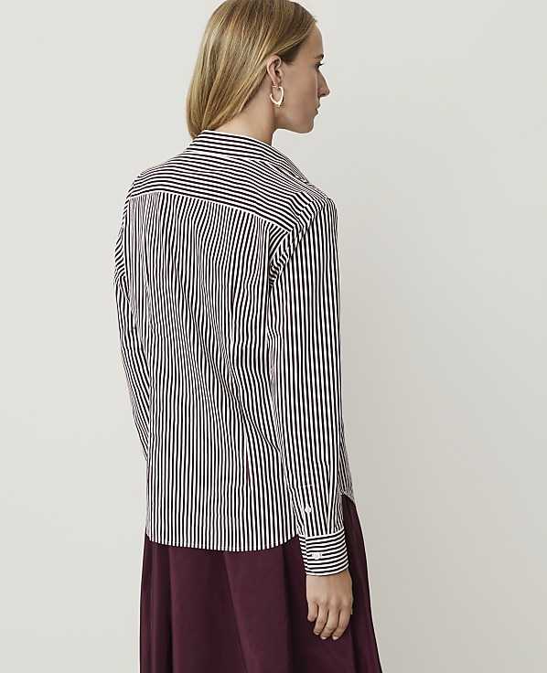 A woman in a striped shirt and maroon skirt stands against a plain white background.