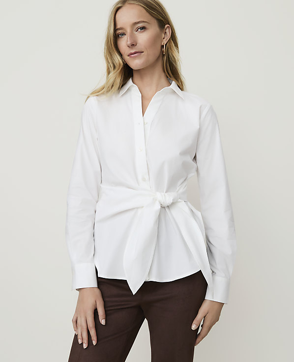 A woman in a white tie-front blouse and brown pants stands against a plain white background.