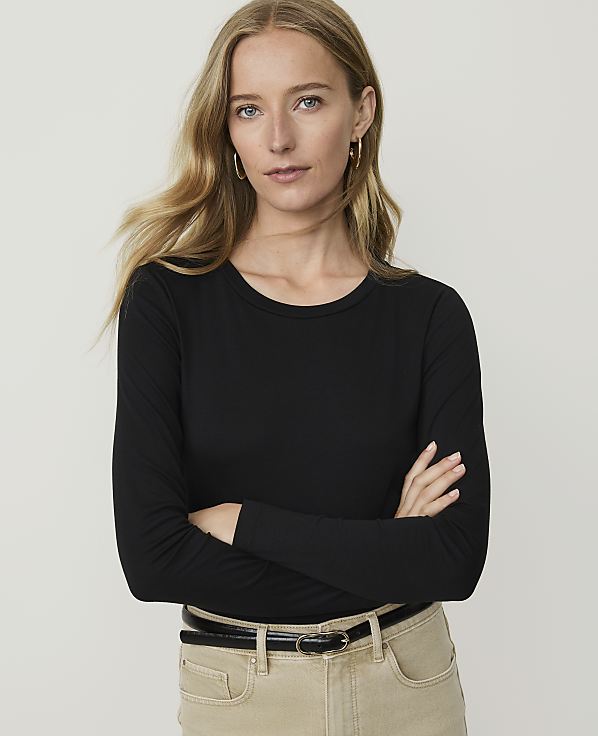 A woman with blonde hair and hoop earrings, wearing a black long-sleeved shirt and tan pants, stands with her arms crossed against a plain white background.