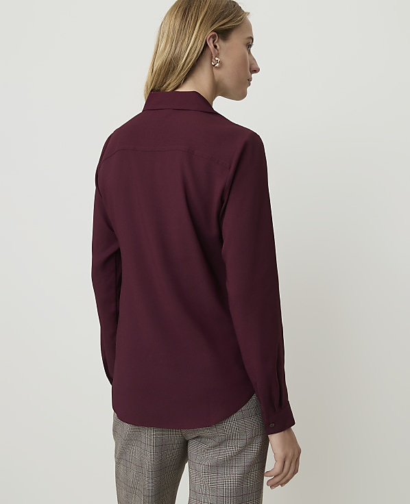 A woman in a maroon collared shirt and plaid pants stands against a plain white background.
