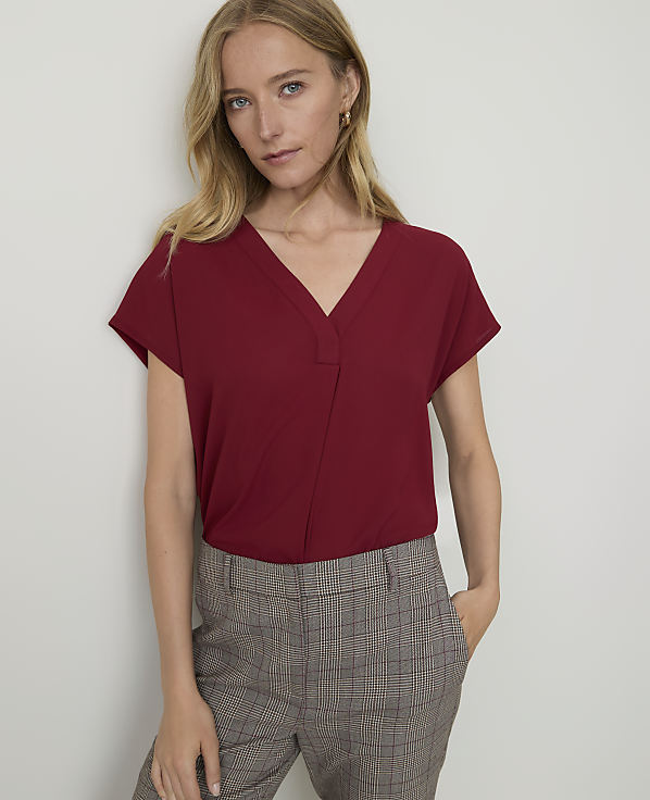 A woman in a maroon v-neck top and plaid pants stands against a plain white wall.