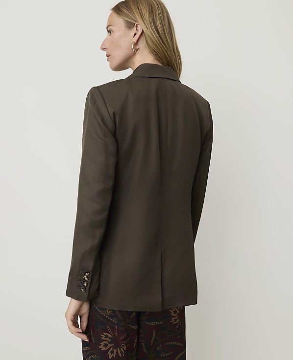 A woman in a brown blazer and patterned pants stands against a plain, light-colored background.