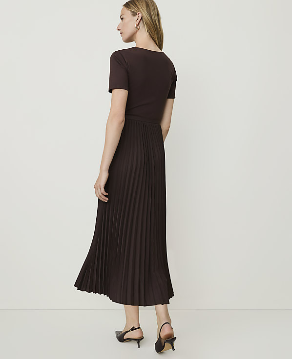 A woman in a brown t-shirt dress with a pleated skirt stands against a plain white background.