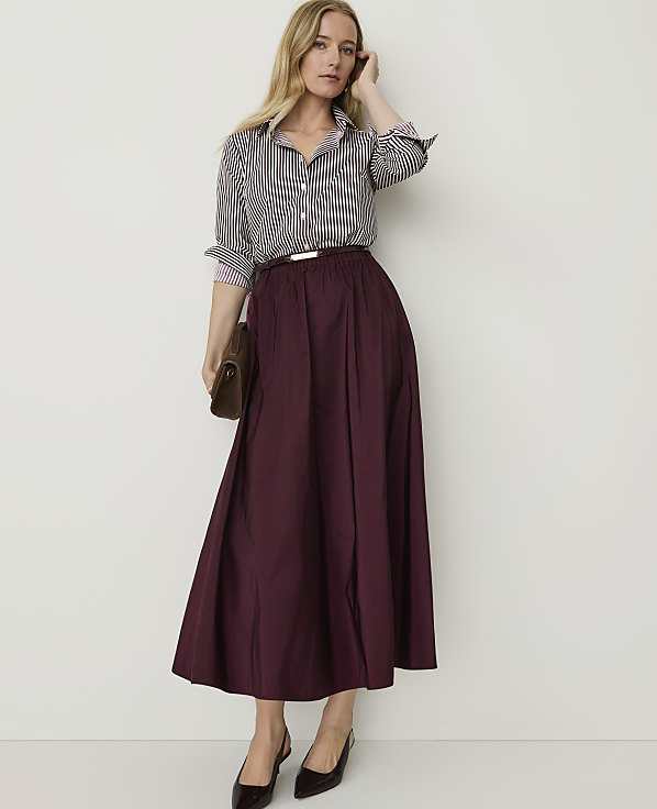 A woman in a striped shirt and burgundy skirt stands against a plain white background, holding a brown clutch.