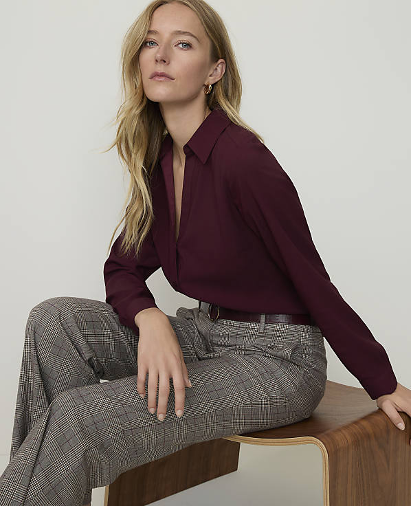 A blonde woman in a burgundy blouse and plaid pants sits on a wooden stool against a plain white background.