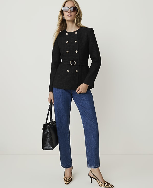 A woman in a black tweed jacket and blue jeans stands against a plain white background, holding a black handbag and wearing leopard print heels.