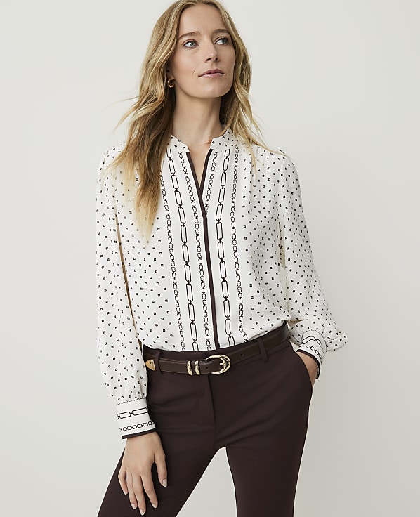 A woman in a patterned blouse and brown pants stands against a plain background.