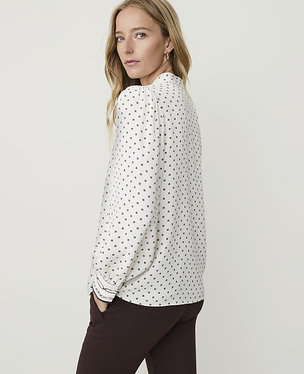 A woman in a white polka-dot blouse and brown pants stands against a plain, light background.