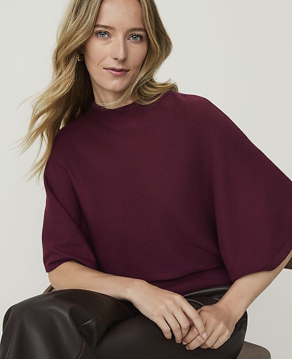 A woman with blonde hair wearing a maroon sweater and dark pants sits in front of a blurred background.