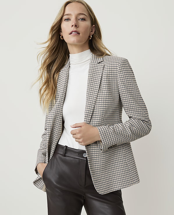 A woman in a checkered blazer and dark pants stands against a plain, light background.