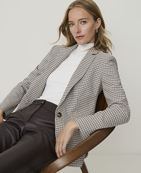 A woman in a checkered blazer and white turtleneck sits in a chair against a plain background.