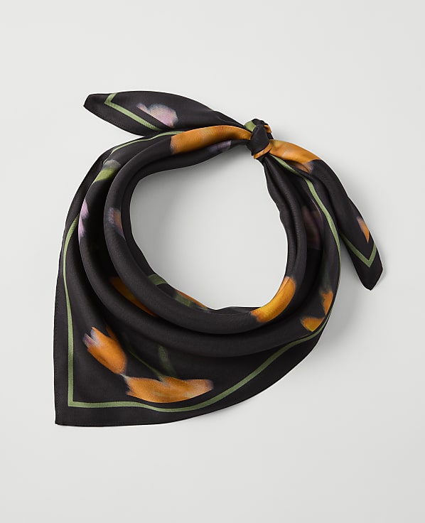 A black silk scarf with an orange floral print and green trim is knotted in the foreground against a plain white background.