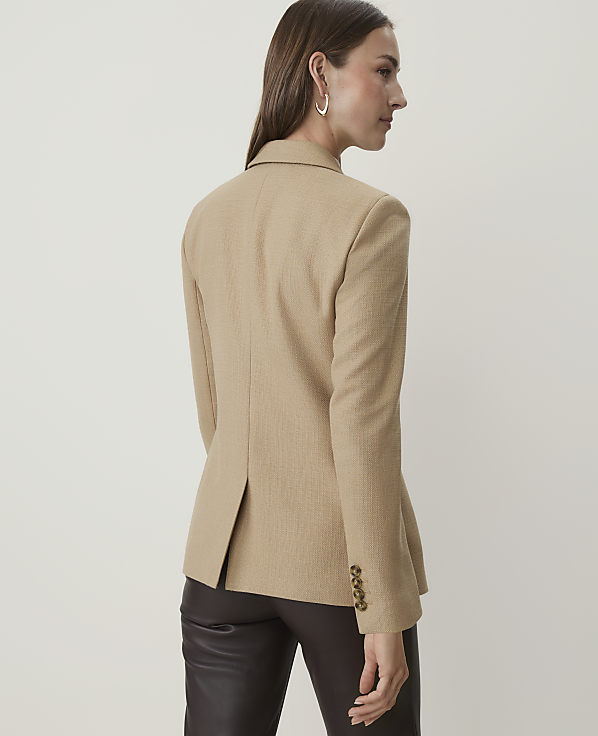 A woman in a tan blazer and dark pants stands against a plain, light-colored background.