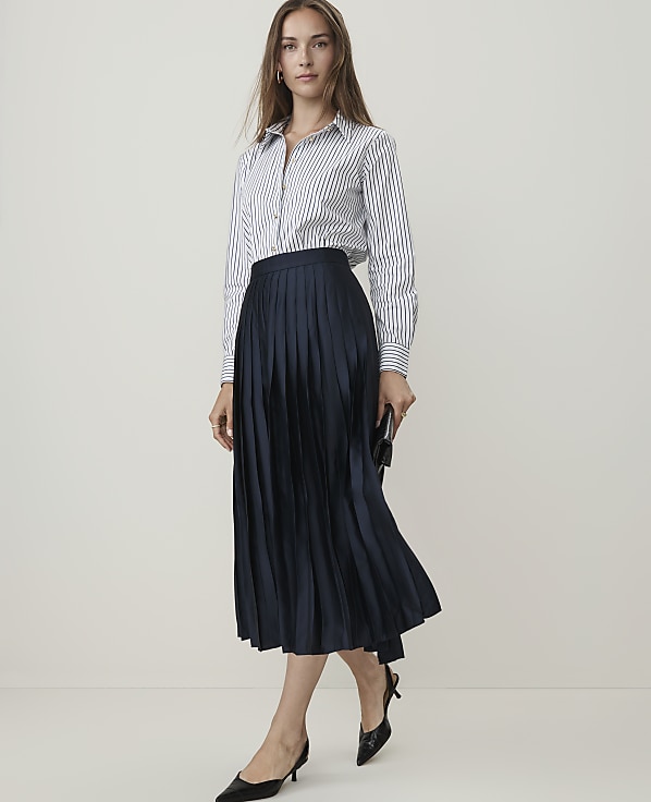 A woman in a striped shirt and navy pleated skirt stands against a plain, light background.