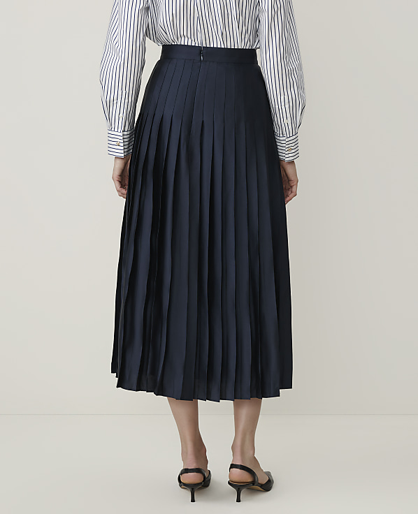 A person stands in the foreground wearing a navy pleated skirt and a striped shirt, against a plain, light-colored background.